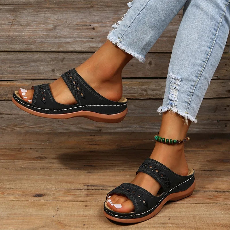 Black woven wedge sandal with double strap and diamond cutout detail, shown on foot against a timber backdrop