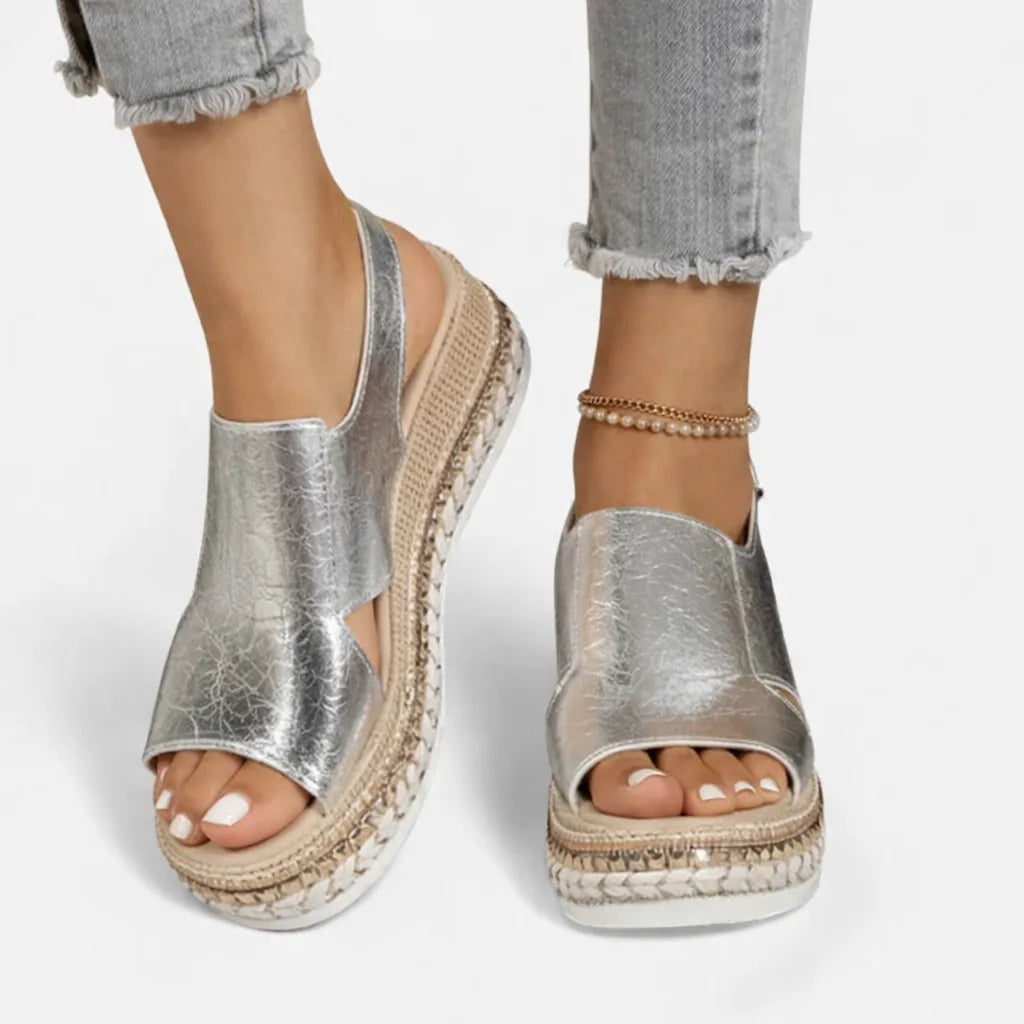 Close-up side view of a silver metallic crinkled-leather wedge sandal with espadrille platform, worn with cuffed grey denim