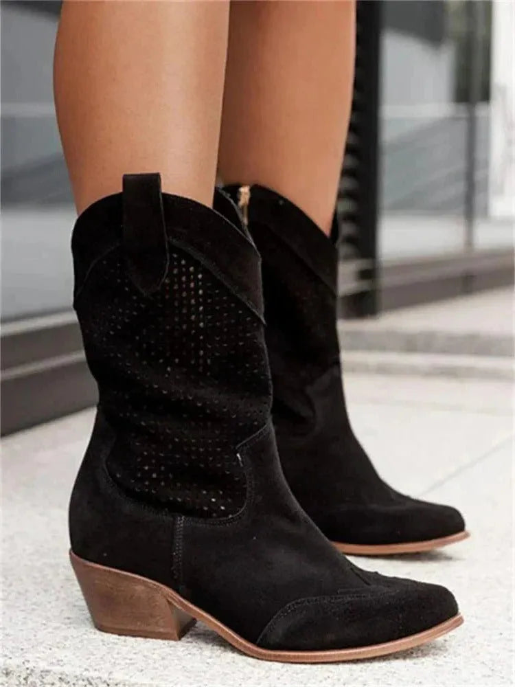 Black suede cowboy boots with laser-cut perforated detailing and wooden block heel, worn on steps outdoors