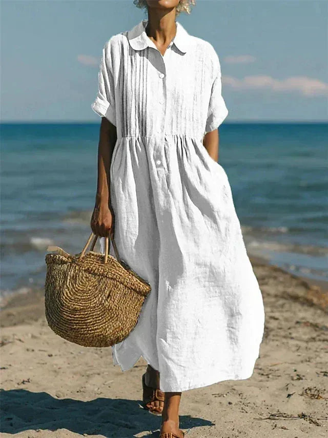 Model walking on a sandy beach in a white pintuck maxi shirt dress paired with a woven straw bag and sandals