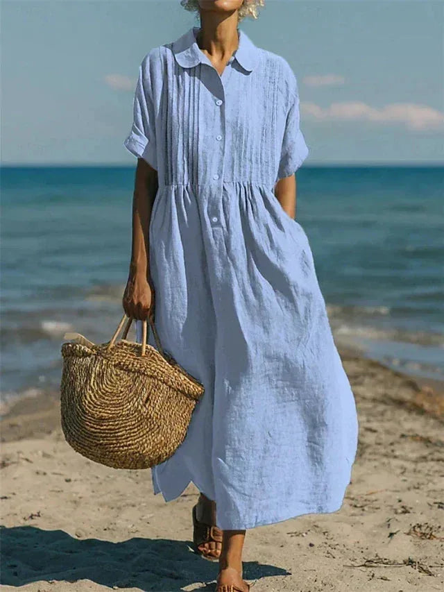 Model on a beach wearing the maxi shirt dress in light blue, carrying a straw tote with the ocean in the background