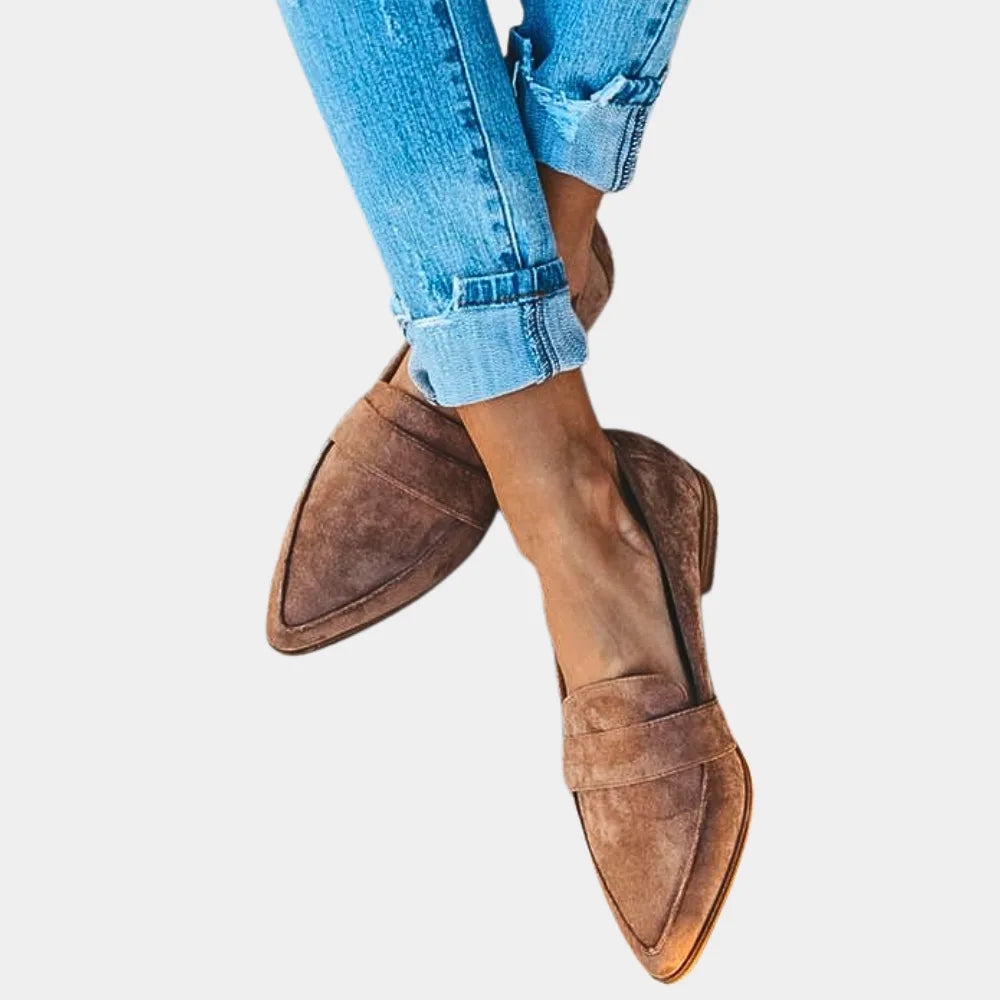 Crossed-leg close-up of brown suede pointed loafers showing the folded vamp strap and stitching