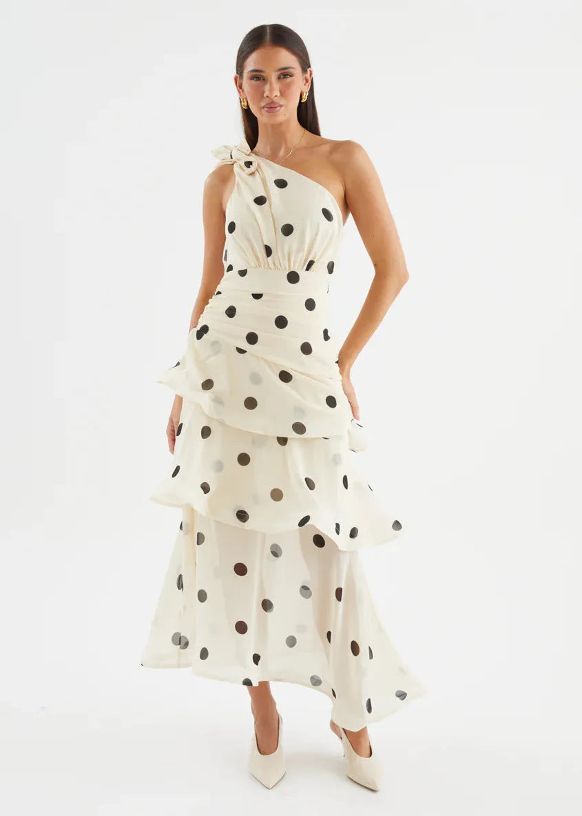 Front view of a model in a cream polka dot one-shoulder midi dress with tiered ruffle skirt and bow detail