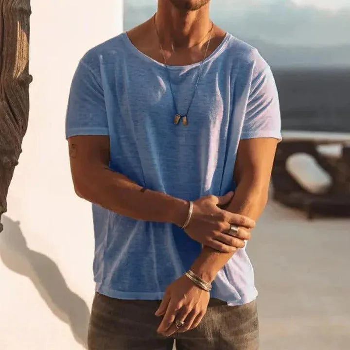 Man wearing a light blue crew neck T-shirt with a relaxed drape, styled outdoors near a white wall and sea view.