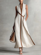 Cream sleeveless maxi dress with contrast tan piping and tie-waist belt, shown with a brown and white tote
