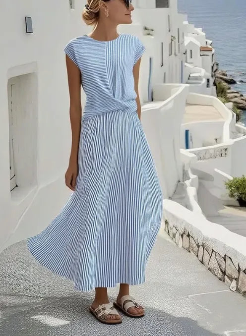 Woman in a blue and white striped maxi dress with twisted waist detail, walking along a sunlit Mediterranean-style terrace.
