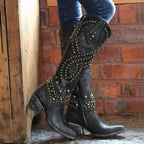 Close-up of black leather studded knee-high cowboy boots worn with jeans against a brick wall