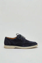 Side profile of navy suede lace-up shoe with moc-toe stitching and off-white sole on a grey background