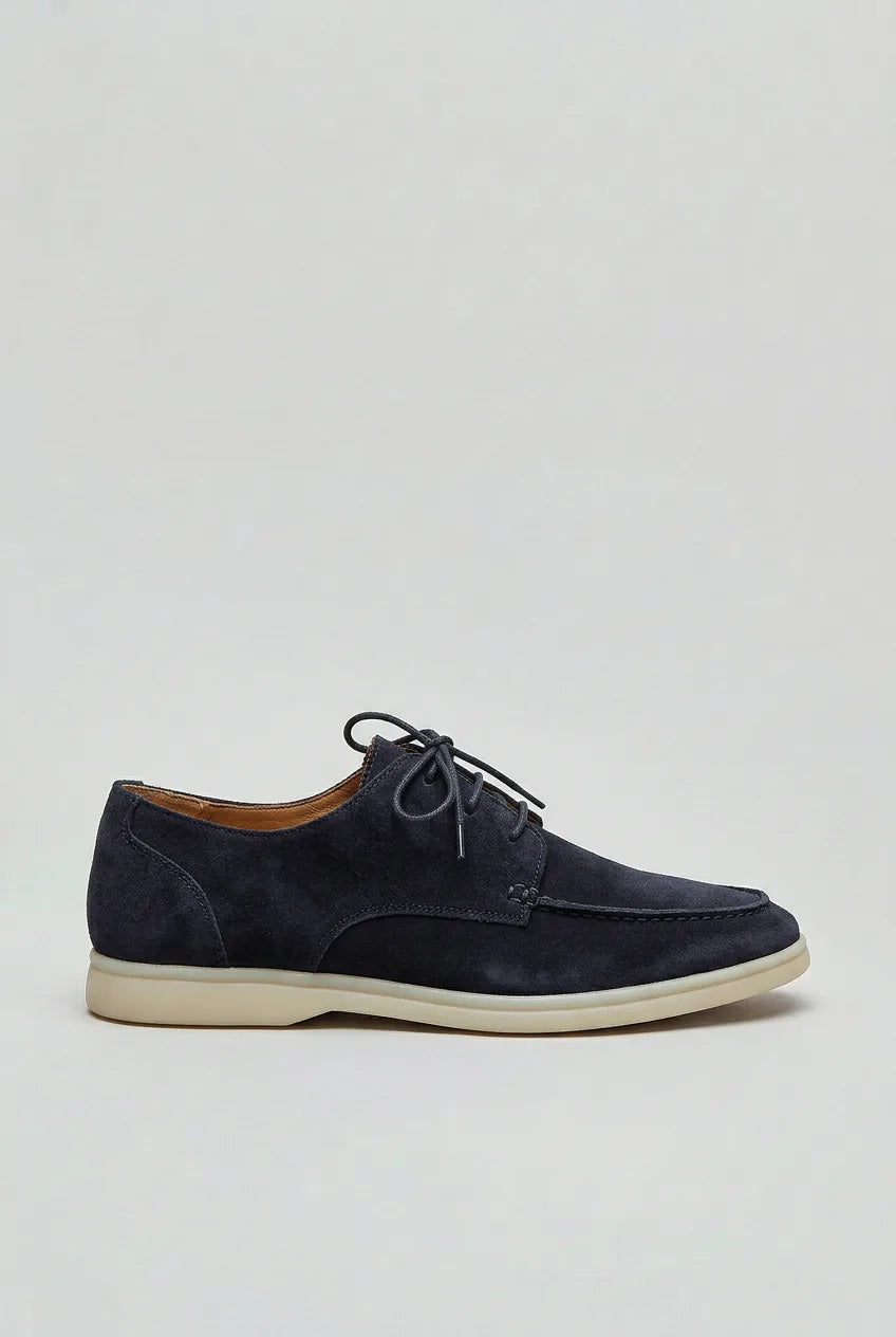 Side profile of navy suede lace-up shoe with moc-toe stitching and off-white sole on a grey background