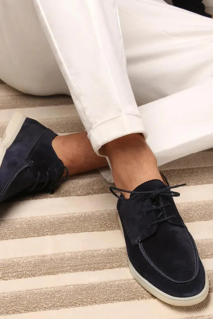 Man seated wearing navy suede lace-up shoes with white cuffed trousers, close-up on a striped rug