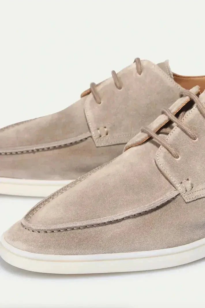 Close-up front view of beige suede lace-up shoes highlighting the stitched apron toe and laces