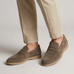 Close-up side view of taupe suede penny loafers worn with cuffed beige linen trousers, showing gum sole detail