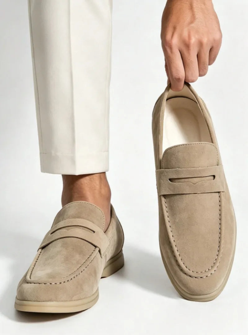 Close-up side view of beige suede penny loafer held by hand, worn with cream tailored trousers