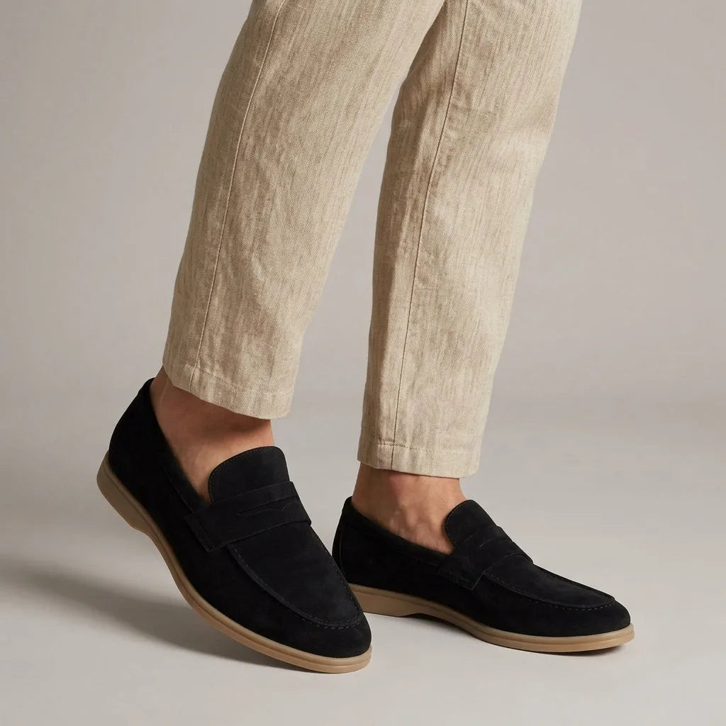 Angled view of navy black suede penny loafers on feet, paired with light beige linen pants against a grey backdrop