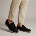 Angled view of navy black suede penny loafers on feet, paired with light beige linen pants against a grey backdrop