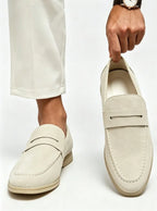 Off-white suede penny loafer being lifted by hand, paired with cream cropped trousers and a watch