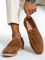 Tan brown suede penny loafer held up by hand, shown next to a bare ankle and cream trousers