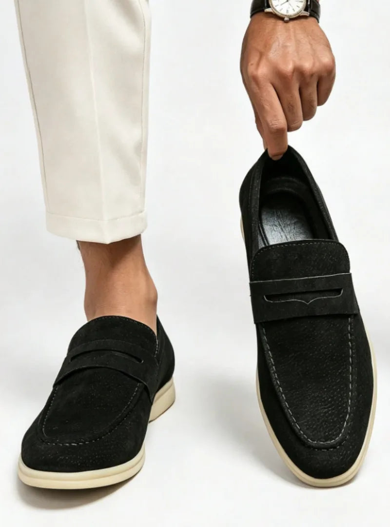 Black suede penny loafer with contrast cream sole, pinched by hand above a matching worn shoe