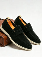 Pair of black suede penny loafers with cream soles and wooden shoe trees beside a leather bag