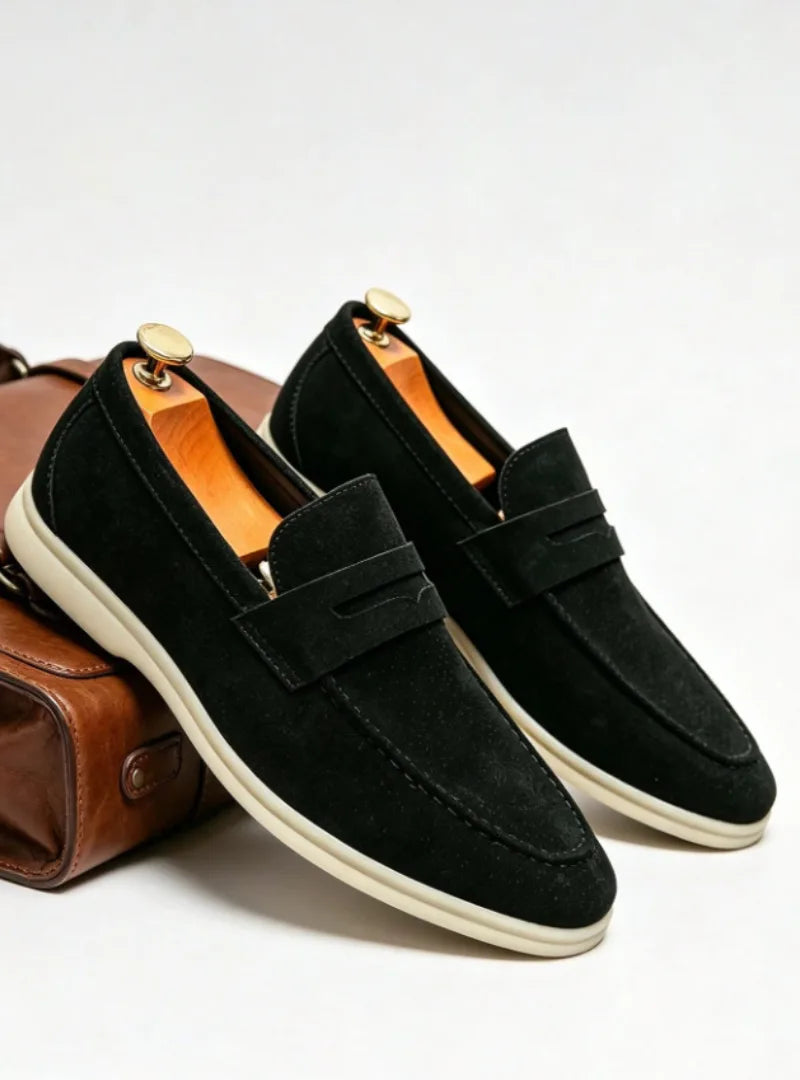 Pair of black suede penny loafers with cream soles and wooden shoe trees beside a leather bag