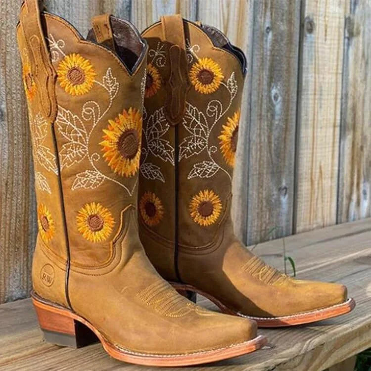 Tan leather cowboy boots with sunflower and vine embroidery on the shaft, paired against a timber fence backdrop.
