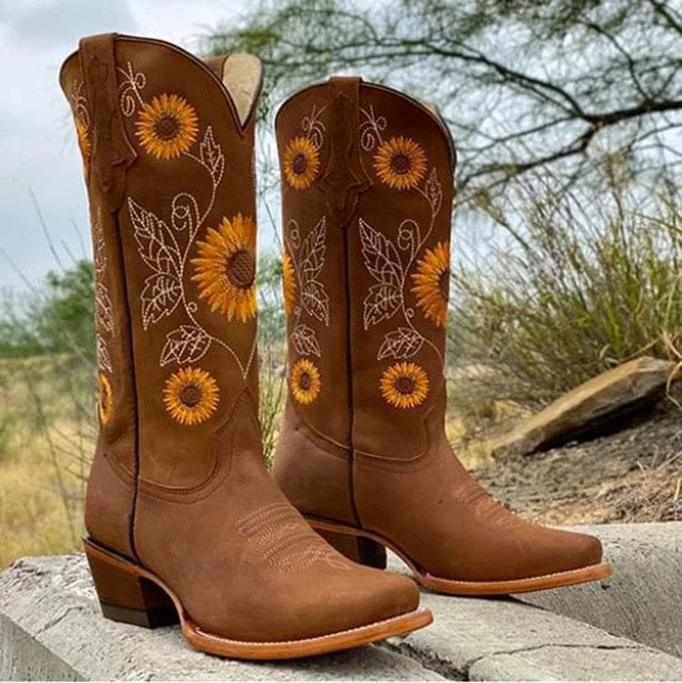Light brown cowboy boots with floral sunflower embroidery and a stacked block heel, standing outdoors on a stone ledge.