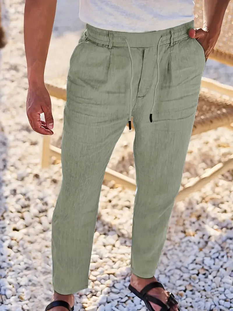 Man standing on pebbled ground wearing sage green tapered linen pants with corded drawstring waistband