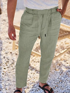 Man standing on pebbled ground wearing sage green tapered linen pants with corded drawstring waistband