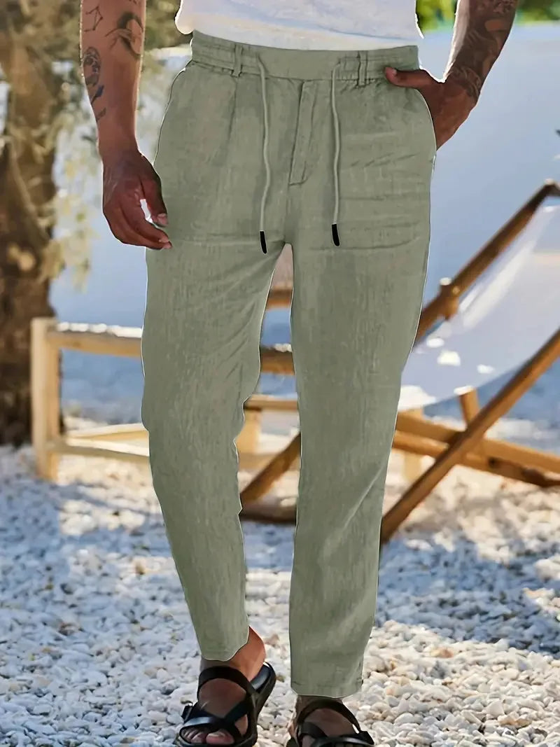 Man outdoors near a lake wearing sage green tapered linen pants with visible pocket stitching and cord tie