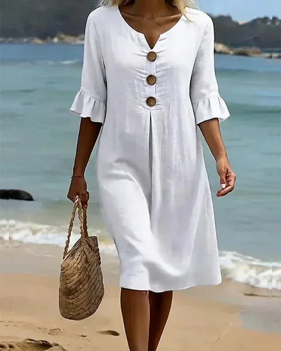 Full-length front view of white shift dress with V-neck and ruffle cuffs, paired with a woven straw tote on the sand