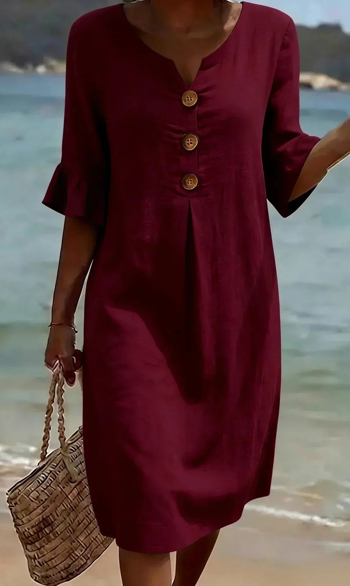 Front view of burgundy textured shift dress with button placket and ruffle cuffs, straw bag in hand