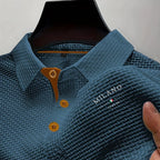 Teal blue textured polo shirt held by hand, showing fabric weave texture and embroidered logo on chest