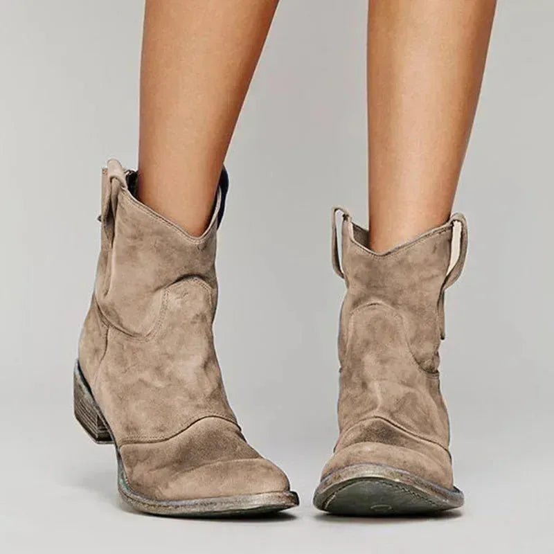 Front view of taupe suede western ankle boots with curved cowboy silhouette and pull tabs, worn on bare legs.