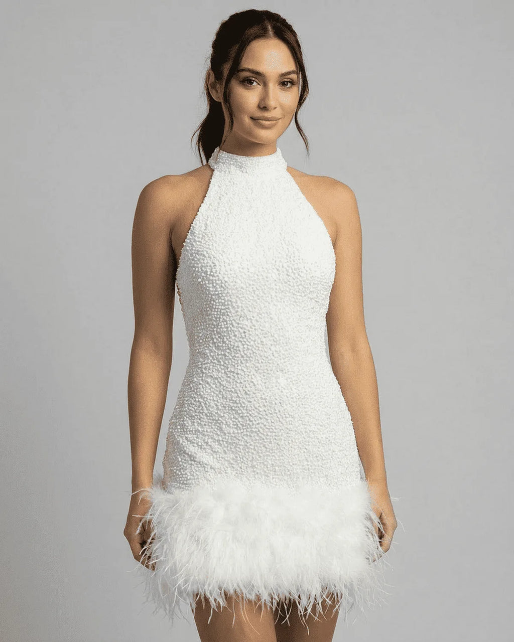 Front view of a model wearing a white sequin halter mini dress with a feathered hem against a grey studio backdrop