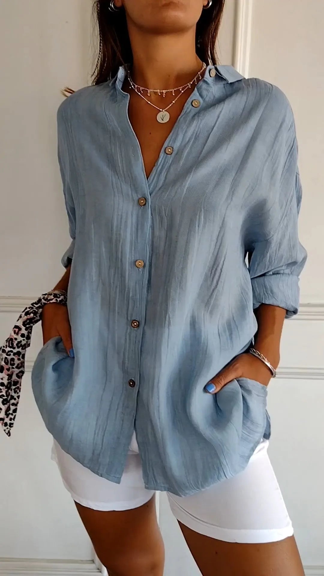 Model wears an oversized sky blue linen shirt with pointed collar, V-neckline and horn-style buttons, worn open over white shorts